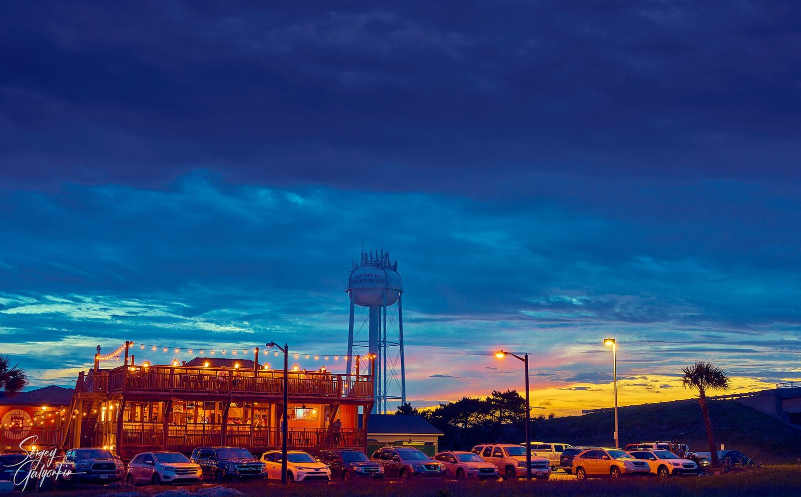 A lit-up waterfront restaurant and the Holden Beach water tower at dusk
